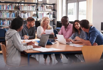 A diverse group of six adults sits around a table in a library or study room, engaged in a collaborative discussion. Laptops, papers, and notebooks are spread across the table as they talk and share ideas. Bookshelves and windows in the background create a bright, academic setting.